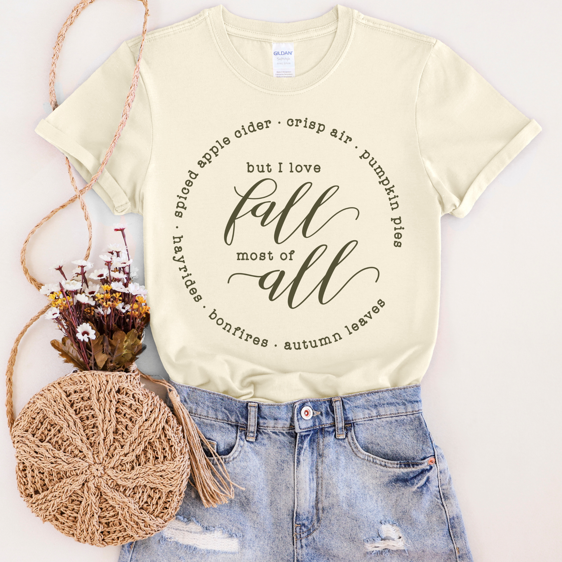 Beige t-shirt with autumn-themed text, paired with denim shorts and a round woven bag on a white background.