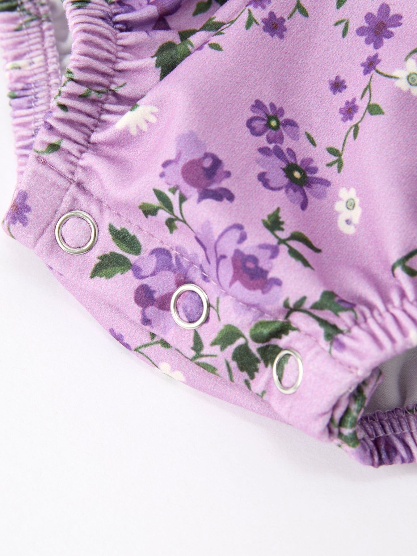 Close-up of a lavender floral garment with purple and white flowers, green leaves, and silver metal snap closures on a white background.