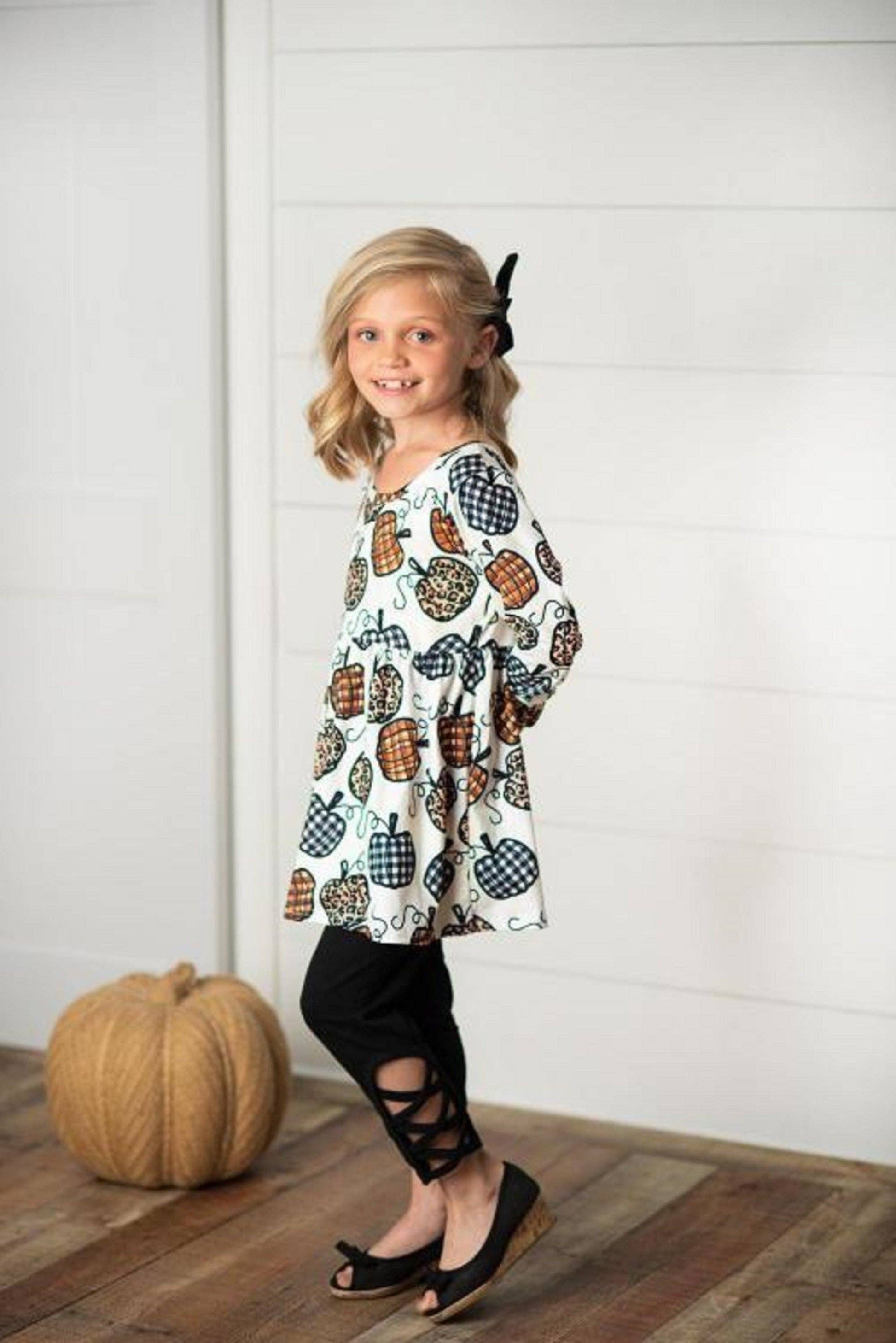 A child standing on a wooden floor wearing a two-piece pant set with a leopard and pumpkin print top and black leggings.