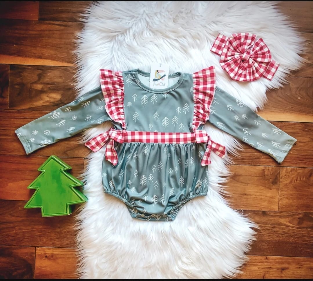 An infant romper with a green color and Christmas themed pattern, laid out on a white furry surface with a red and white checkered blanket and a green Christmas tree ornament beside it.