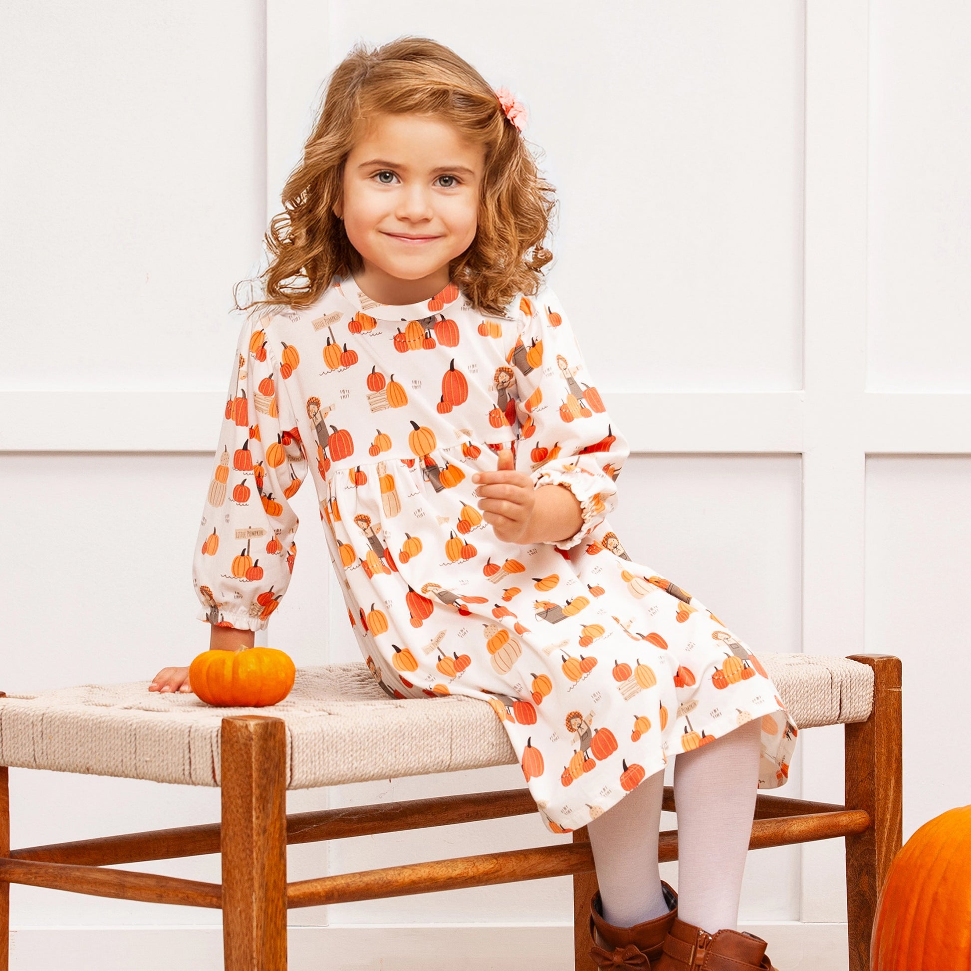 Young girl in a white dress with orange pumpkin pattern sitting on a wooden bench.