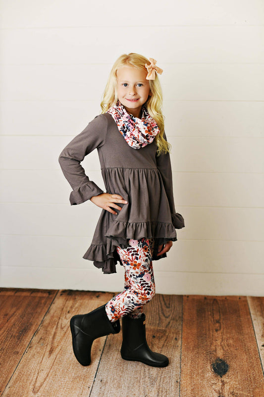 A child model wearing a gray ruffle hi-lo tunic top with long sleeves, paired with multicolored floral print leggings and a matching floral print scarf, standing against a plain background.