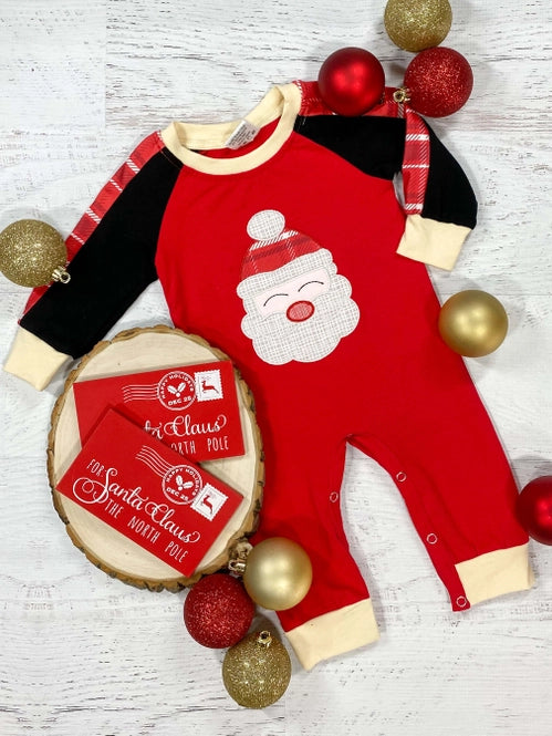 A red one-piece baby romper with a Santa Claus design, displayed with Christmas decorations around it.