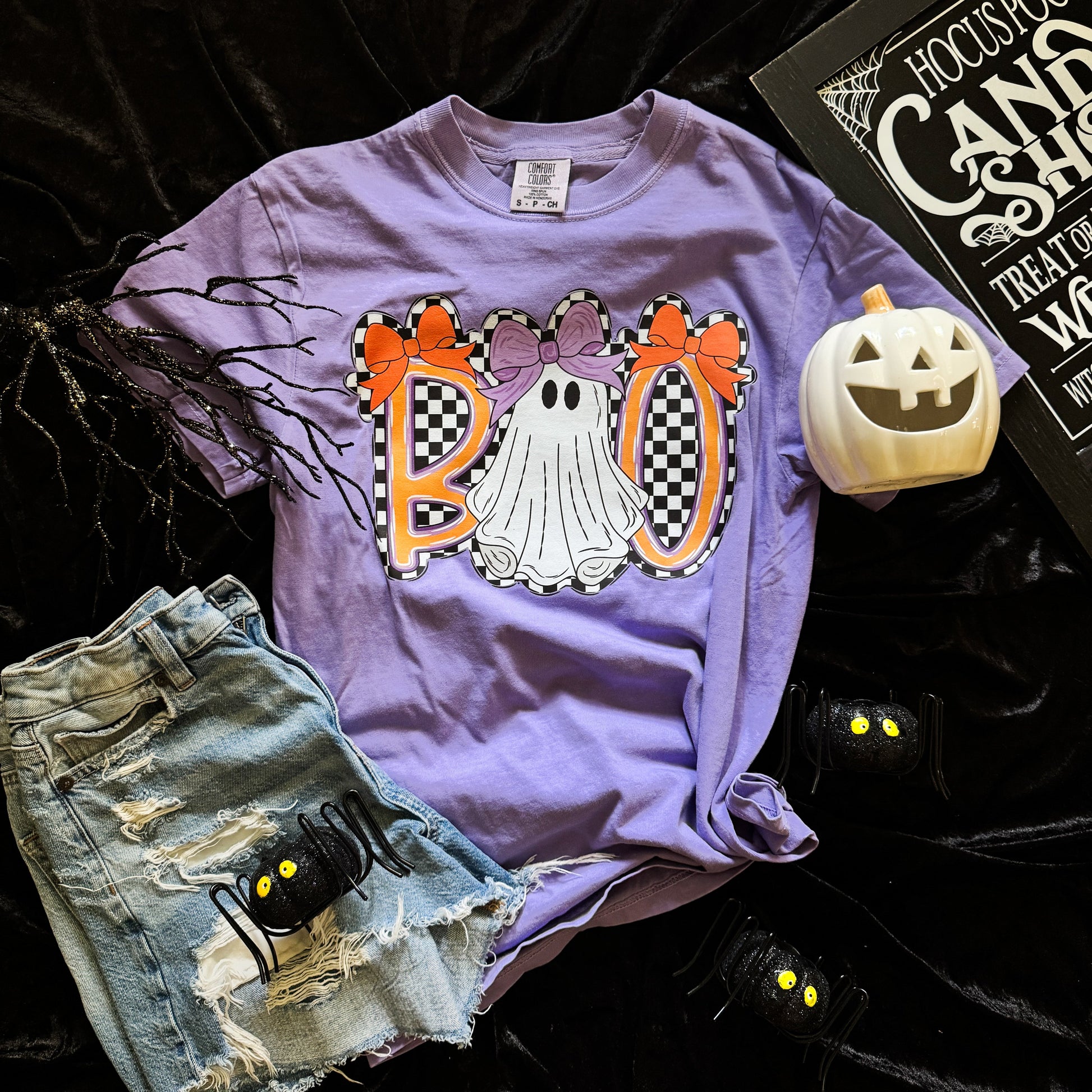 Purple t-shirt with Halloween-themed design featuring ghosts and bows, paired with denim shorts and a small pumpkin on a dark background.