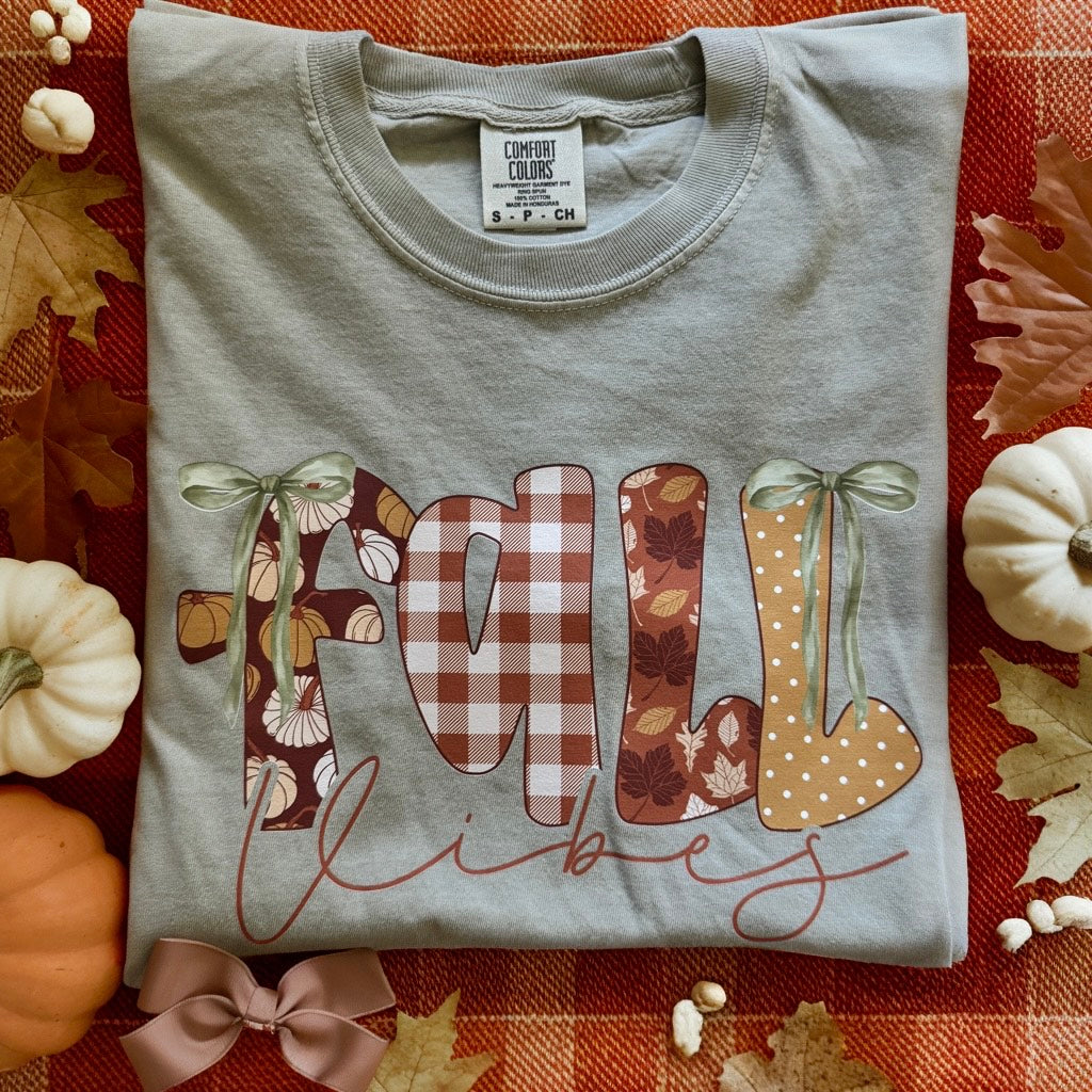 Folded gray t-shirt with 'Fall Vibes' design on a red checkered fabric background with pumpkins and leaves.