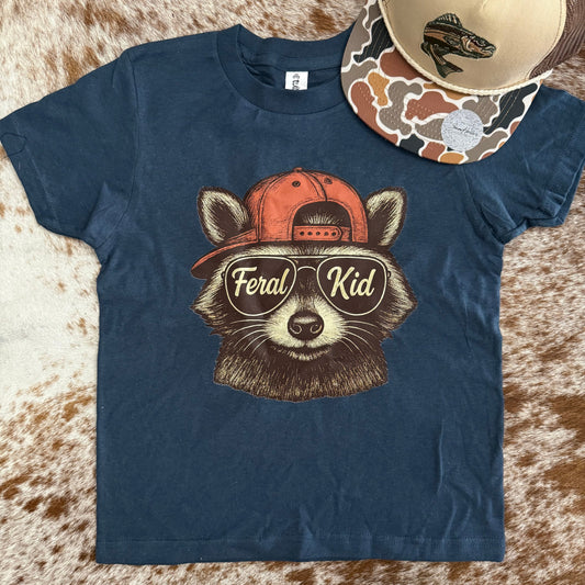 Navy blue t-shirt with a raccoon graphic and 'Feral Kid' text, placed on a textured surface.