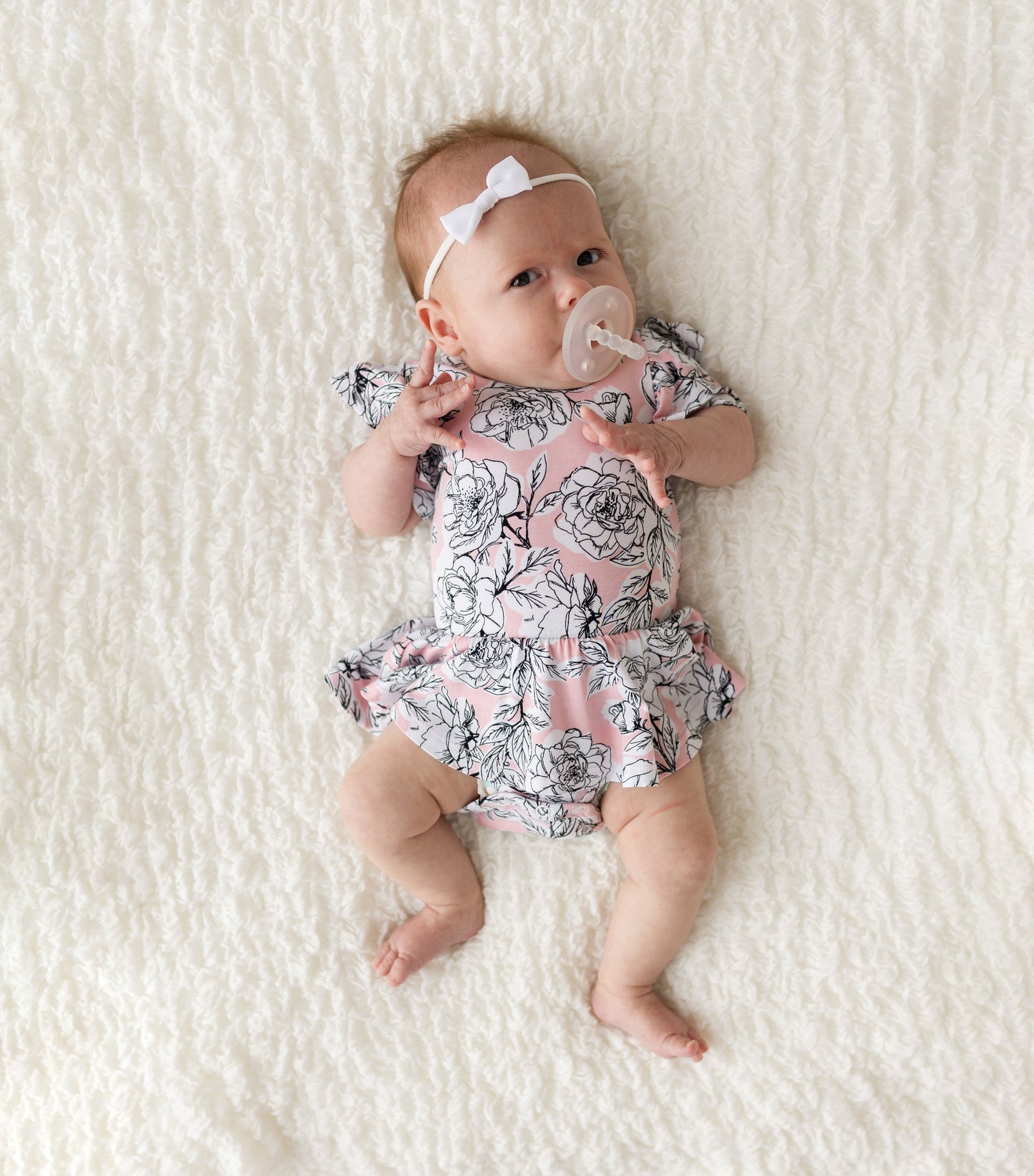 Baby in a floral romper with a pacifier on a white blanket
