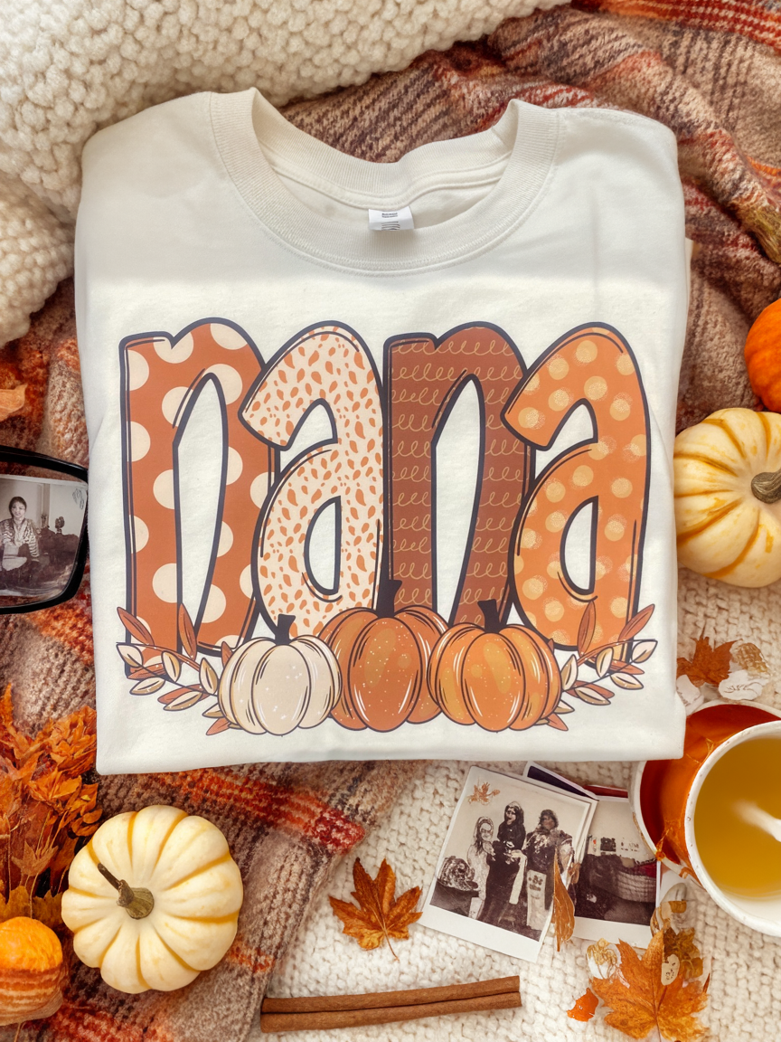 White sweatshirt with 'Nana' and pumpkin design on a fall-themed background with pumpkins and leaves.