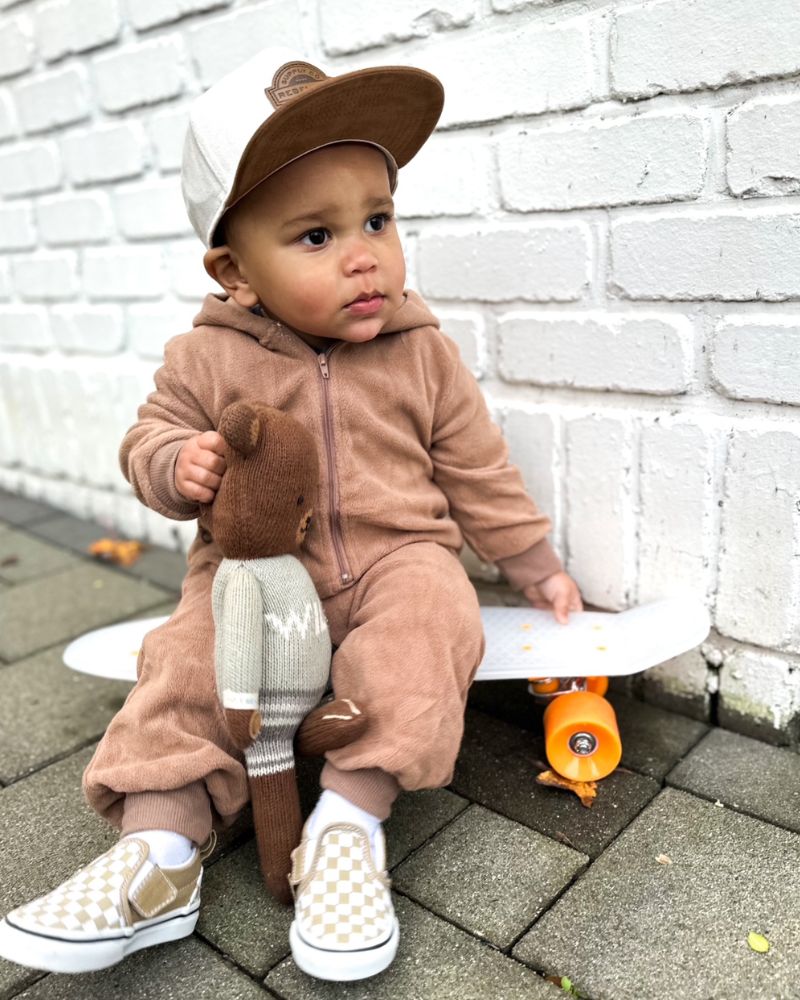 Child in a brown outfit with a bear hat and toy, sitting on a skateboard against a white brick wall.