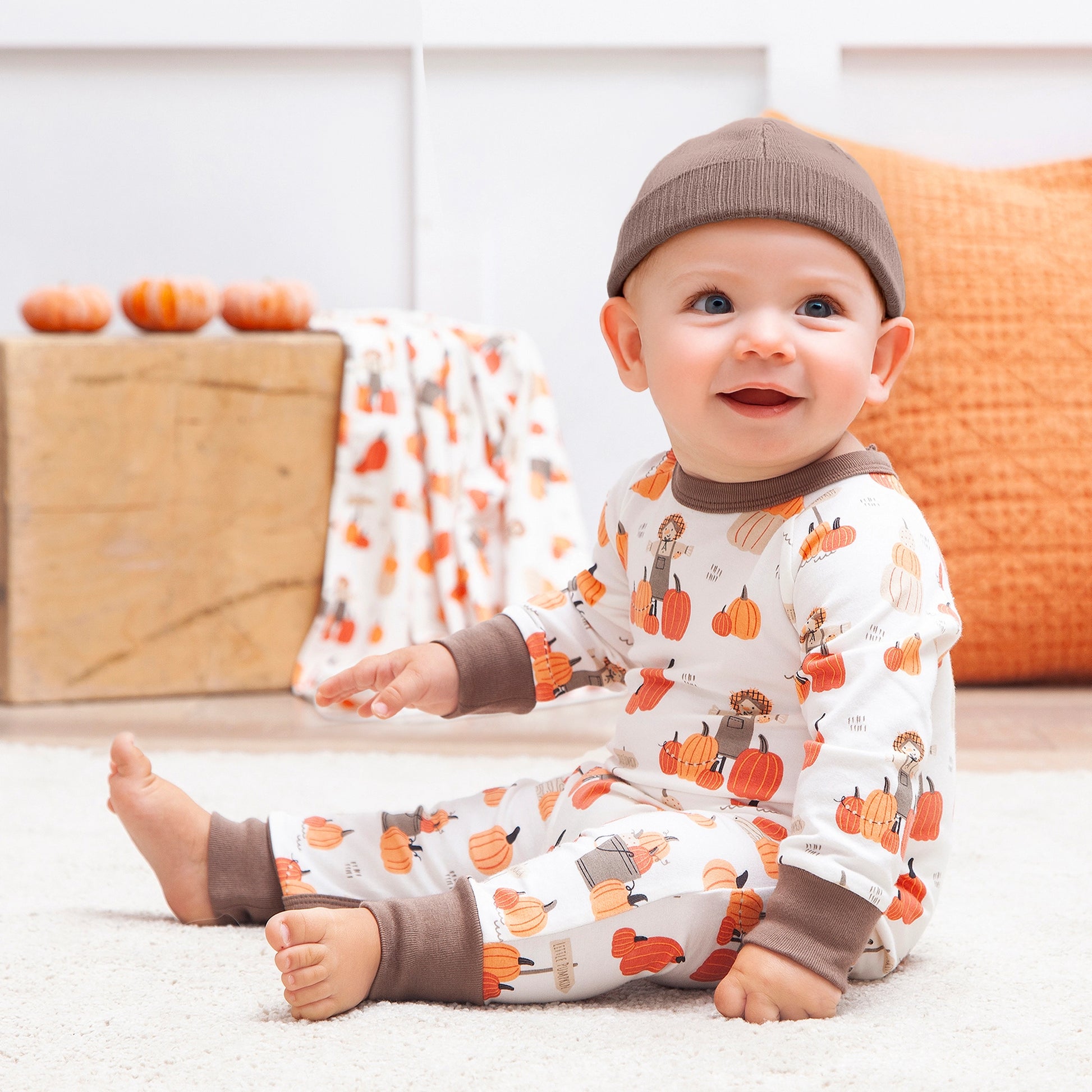 Baby wearing a pumpkin-patterned outfit with a brown hat, sitting on a white surface.
