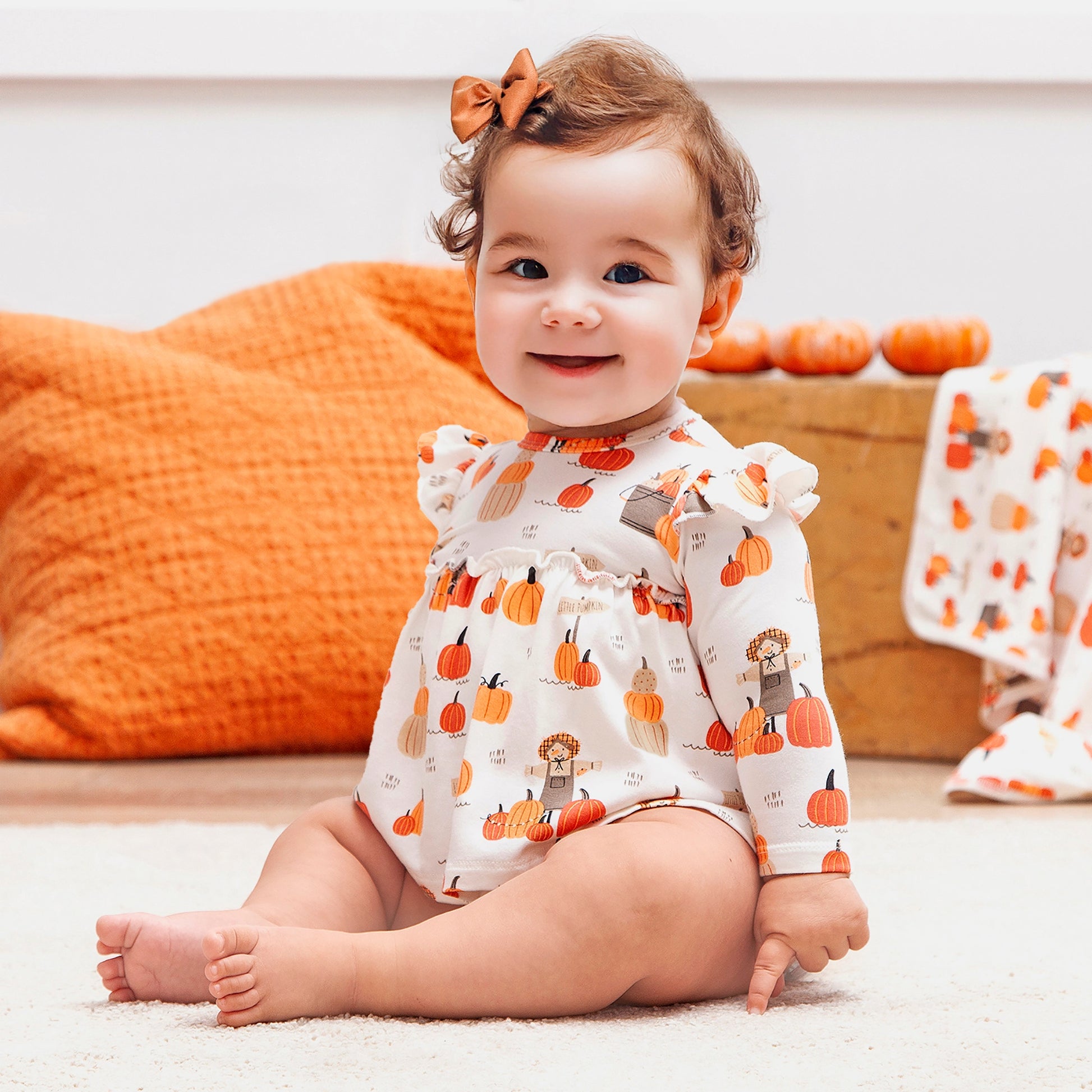Baby wearing a pumpkin-patterned outfit sitting on a carpeted floor with pumpkins in the background.