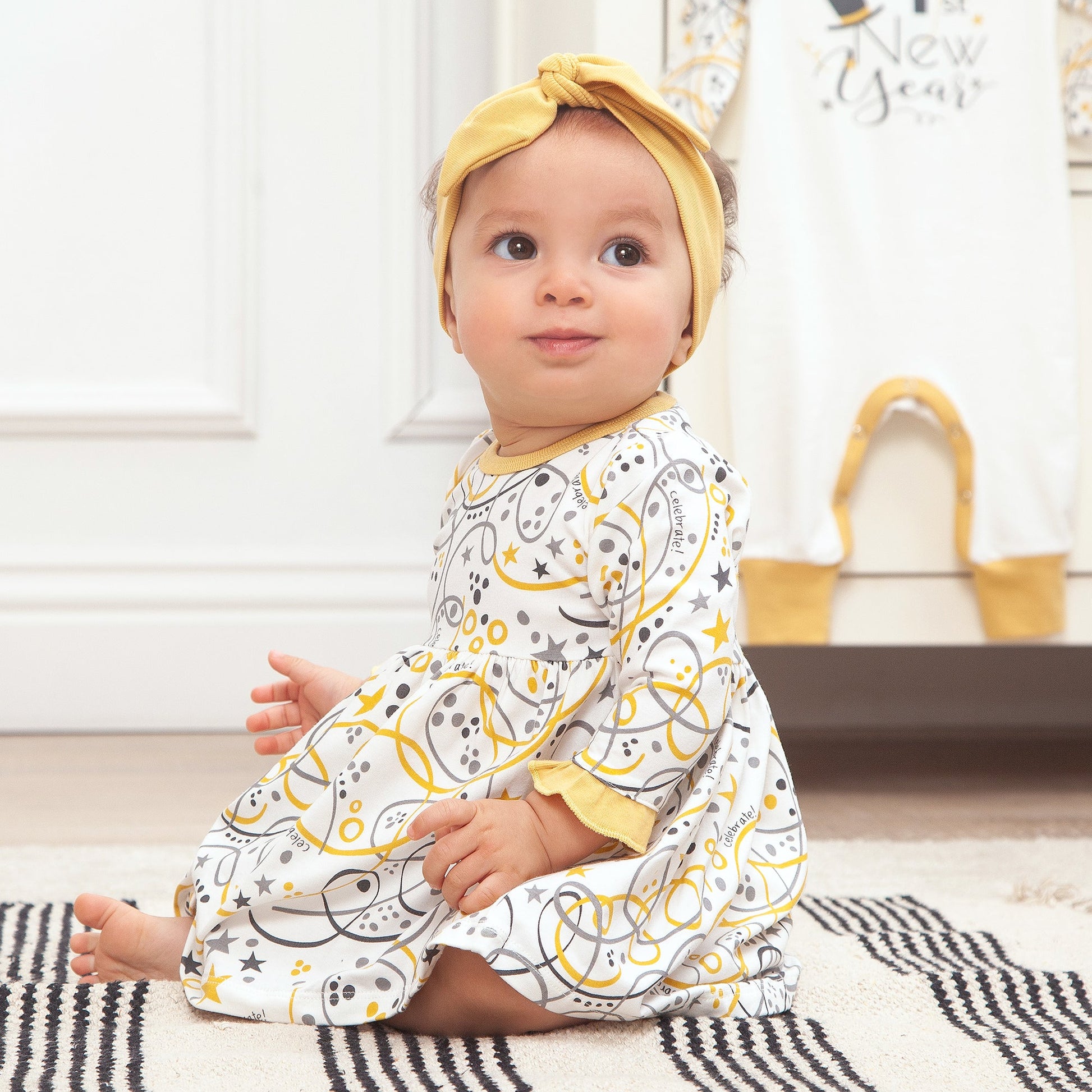 Baby wearing a patterned outfit and yellow headband sitting on a rug.