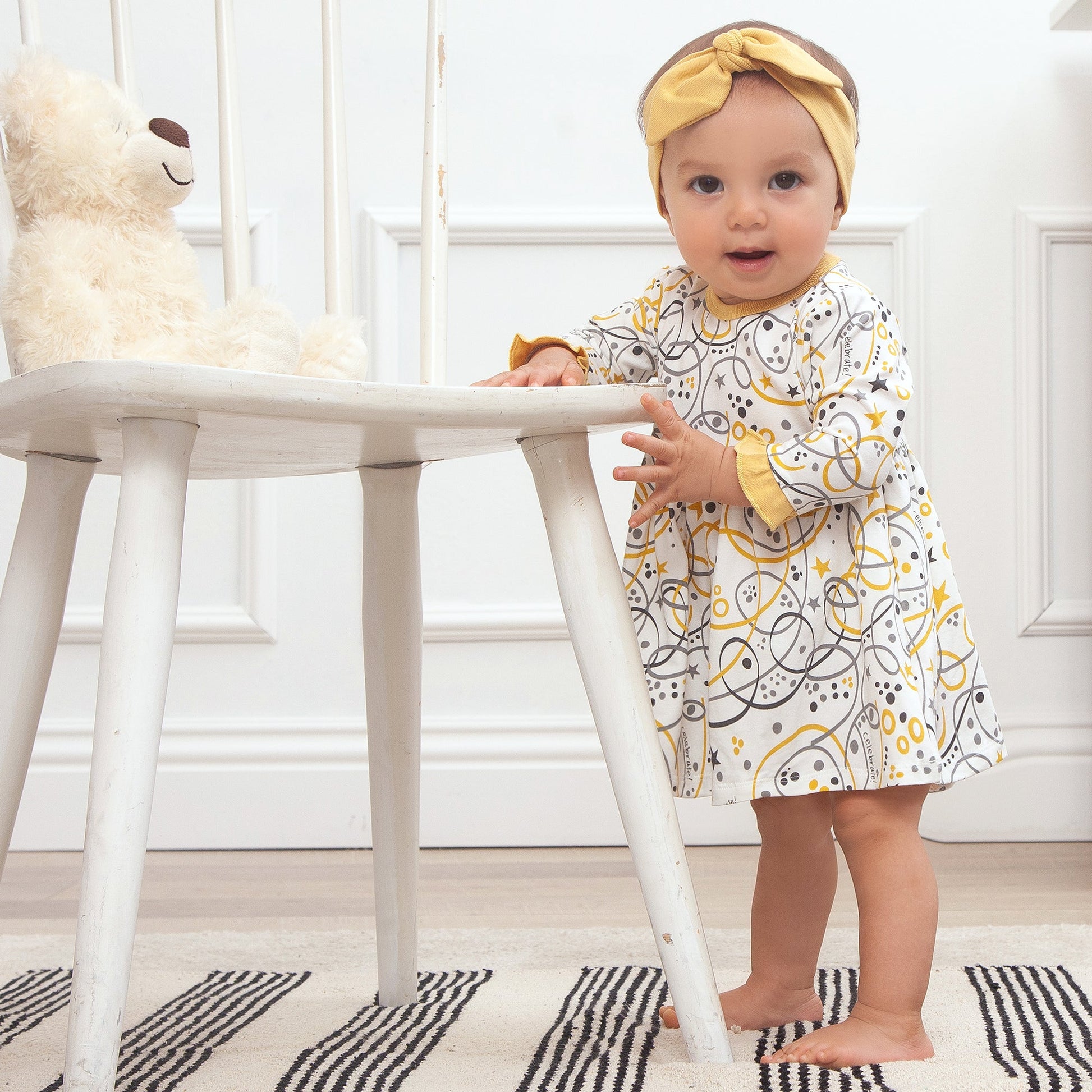 Baby in a white dress with yellow accents standing next to a white table.