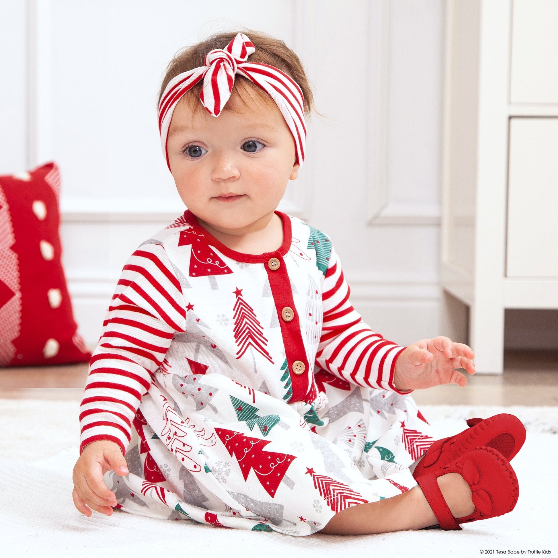 Baby wearing a red and white striped outfit with Christmas tree patterns, sitting on a light-colored floor.