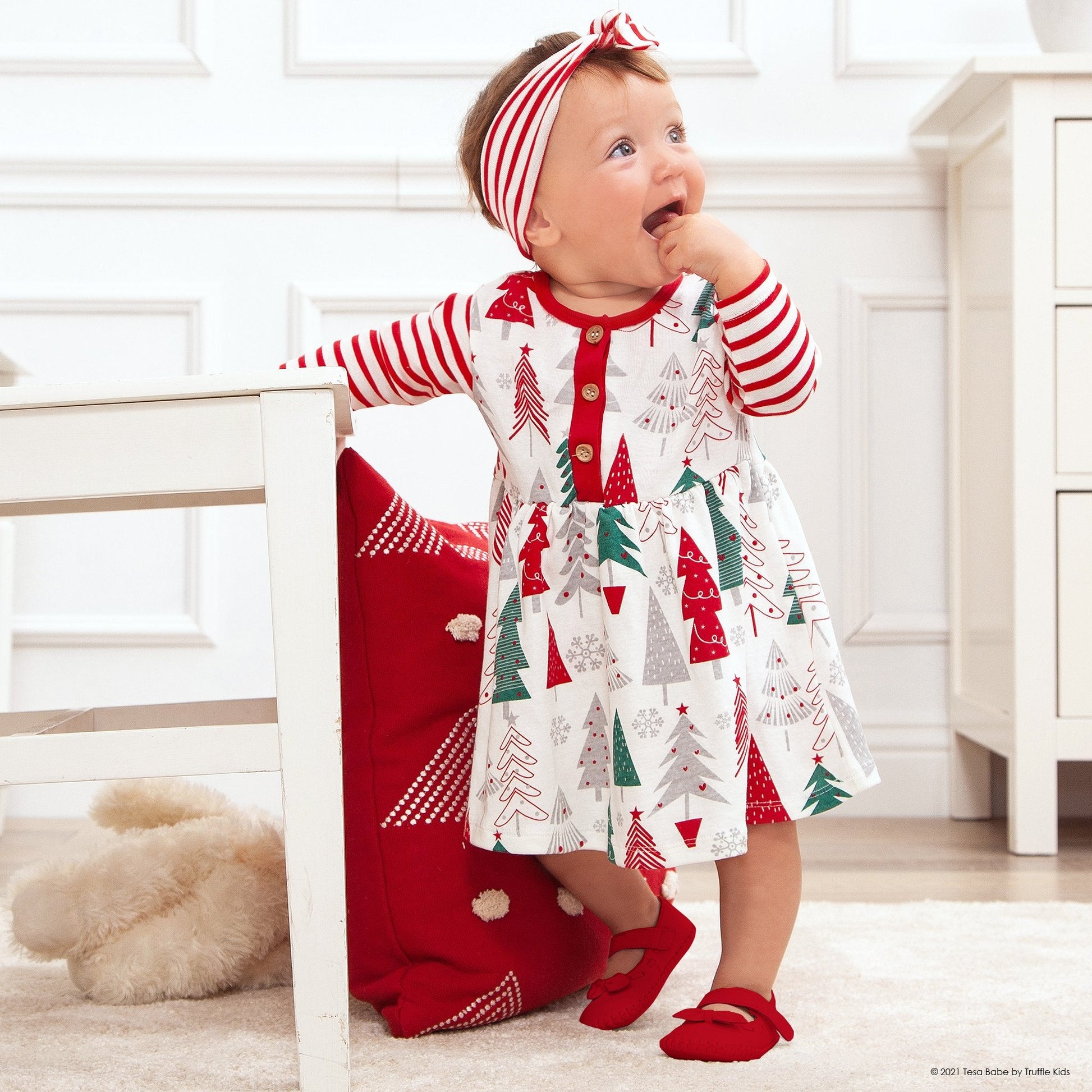 Baby wearing a red and white striped dress with Christmas tree patterns in a home setting.