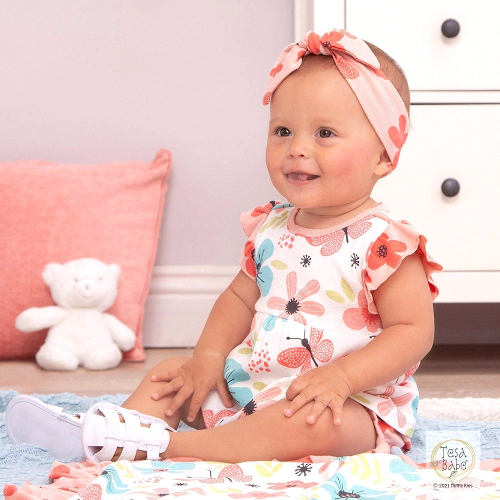 Baby girl in a floral outfit sitting on a bed with a teddy bear and pink pillow in the background.