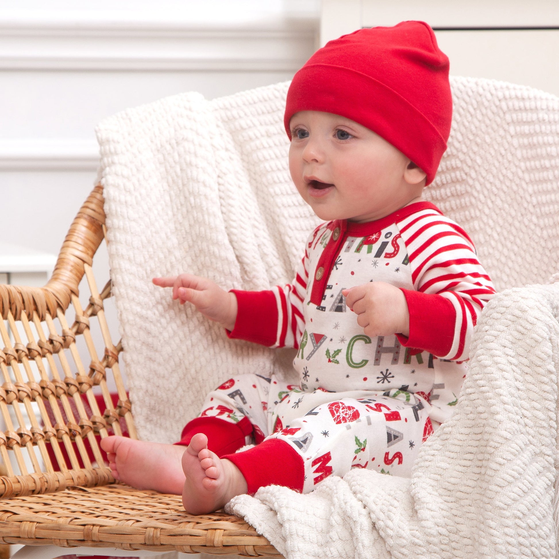 Baby wearing a red hat and Christmas-themed outfit sitting on a wicker chair.
