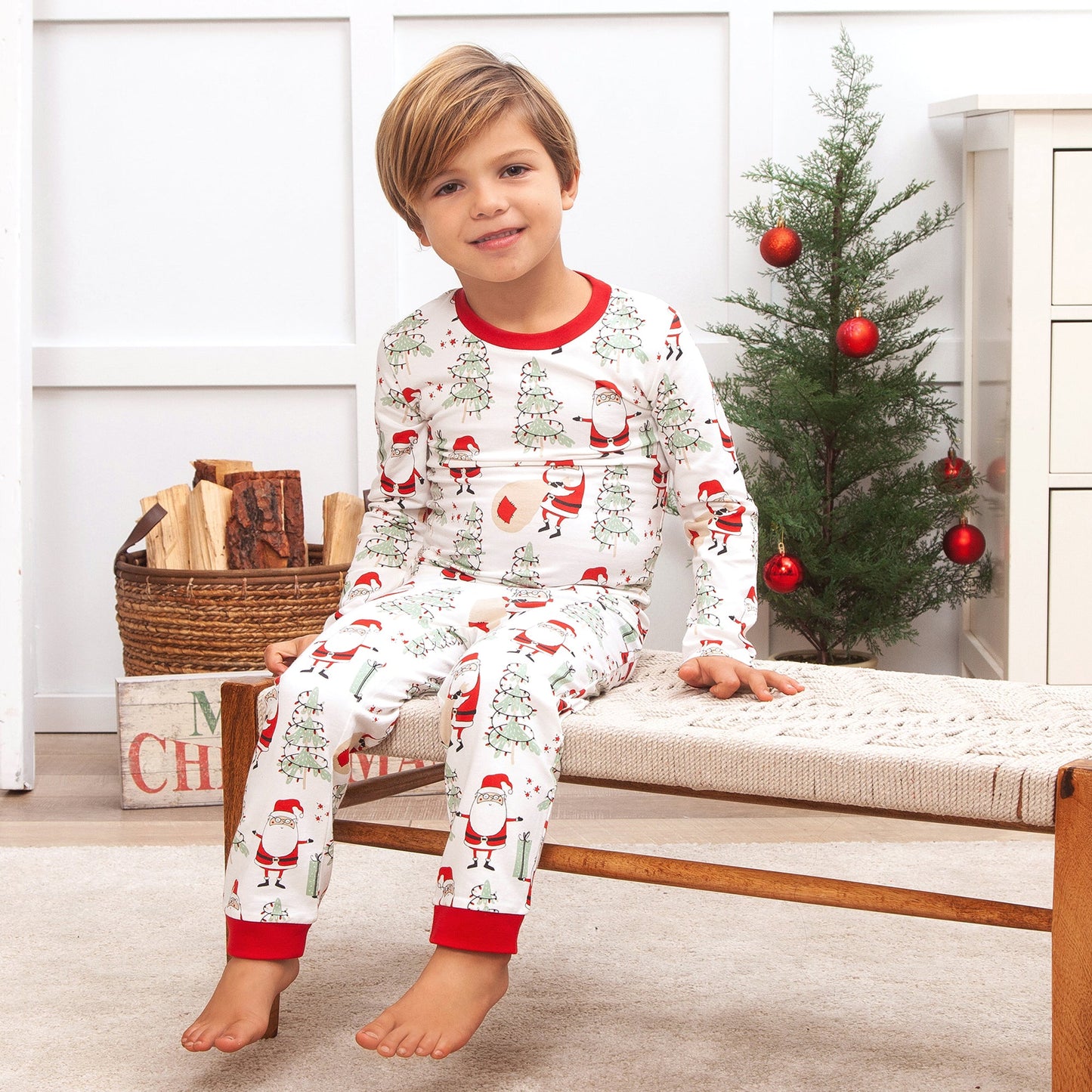 Child wearing a Christmas-themed pajama set sitting on a bench in a room with a decorated tree.