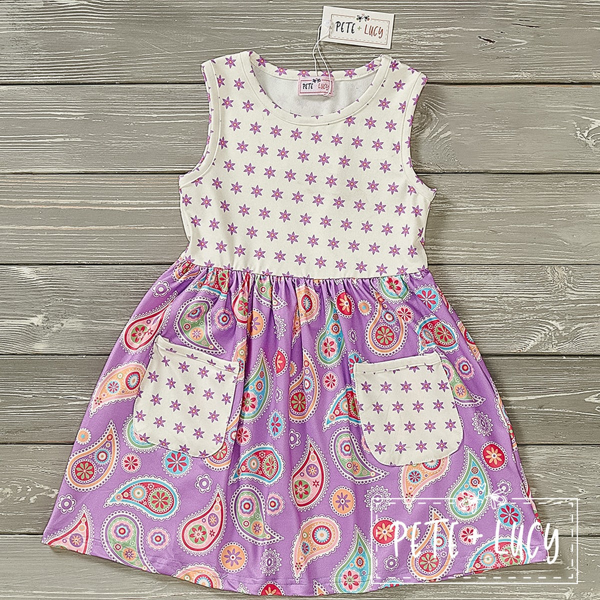 A sleeveless, purple and multicolor paisley patterned dress for kids, with front pockets and a jewel neckline.