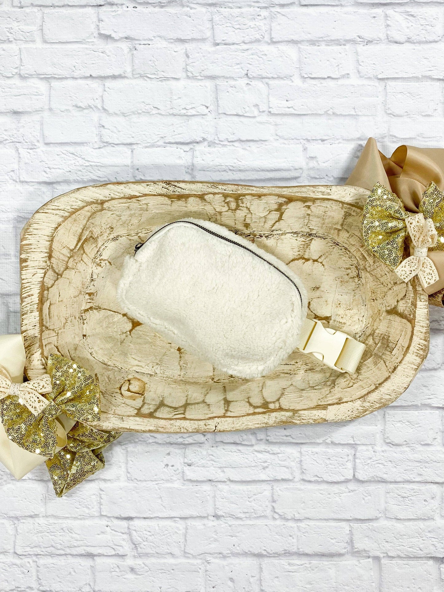 An ivory sherpa belt bag with a textured surface, displayed on a white brick background with decorative gold elements.