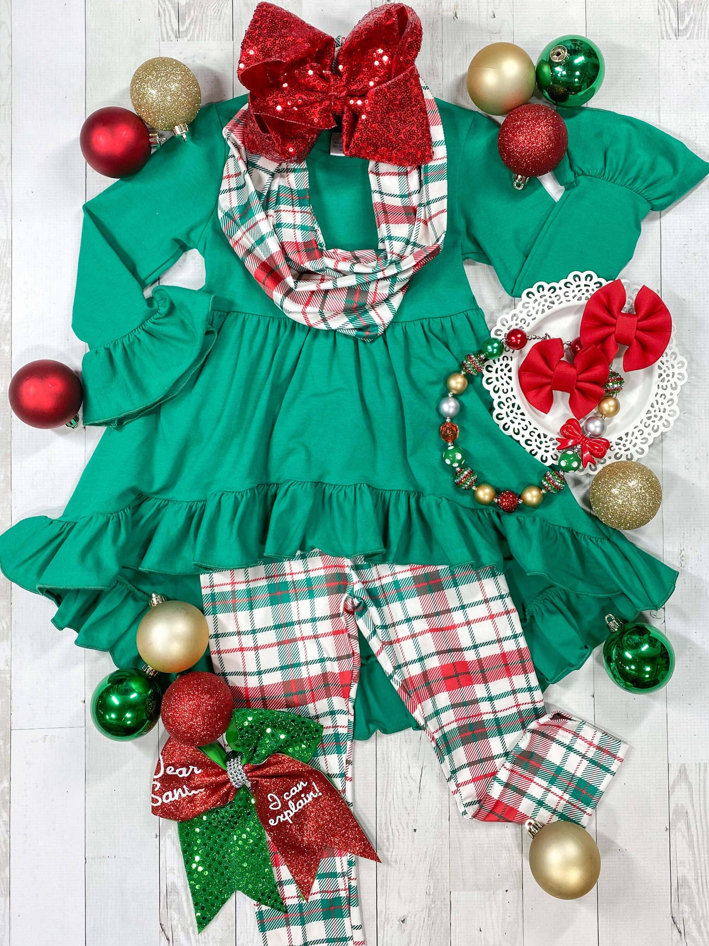 A three piece Christmas plaid set with green and red checkered pattern, including a top with a red bow, pants, and a headband, displayed with Christmas decorations around.