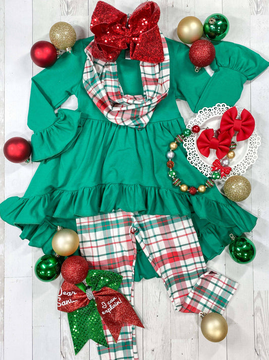 A three piece Christmas plaid set with green and red checkered pattern, including a top with a red bow, pants, and a headband, displayed with Christmas decorations around.