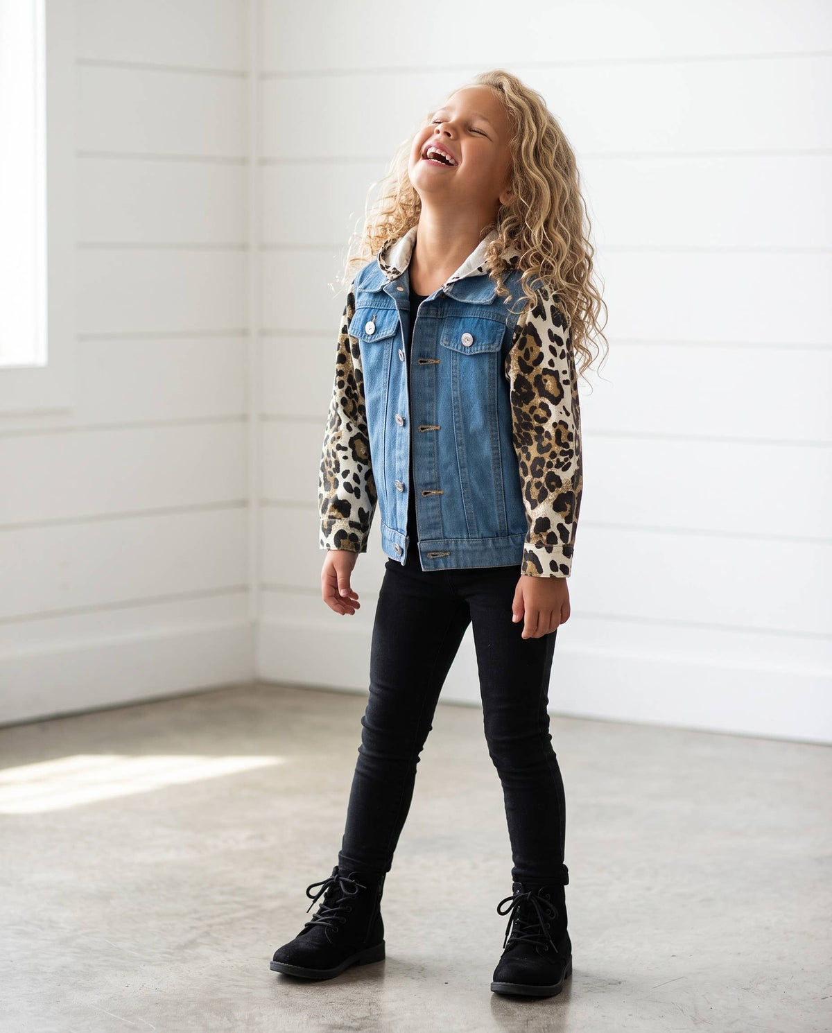 A young girl with curly blonde hair stands in a bright room wearing a denim jacket with leopard-print sleeves, black pants, and black boots.