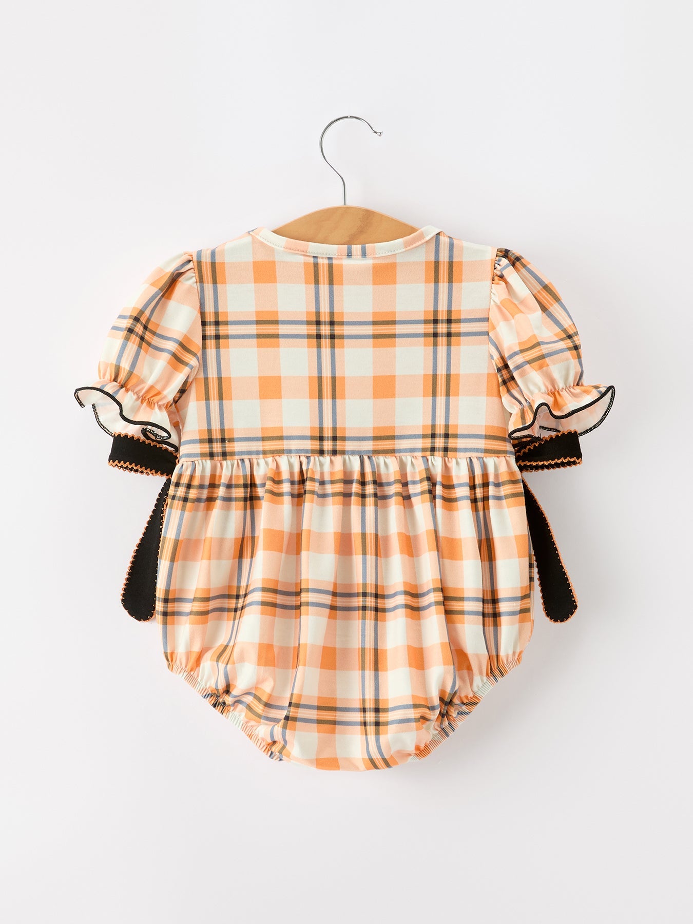 A baby romper with an orange, beige, and black plaid pattern, featuring puff sleeves with ruffled trim and side ties, hanging on a wooden hanger against a plain white background.