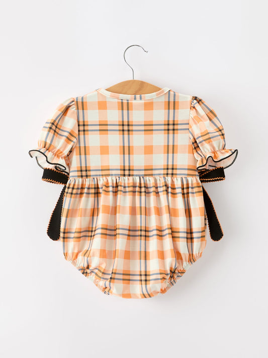 A baby romper with an orange, beige, and black plaid pattern, featuring puff sleeves with ruffled trim and side ties, hanging on a wooden hanger against a plain white background.
