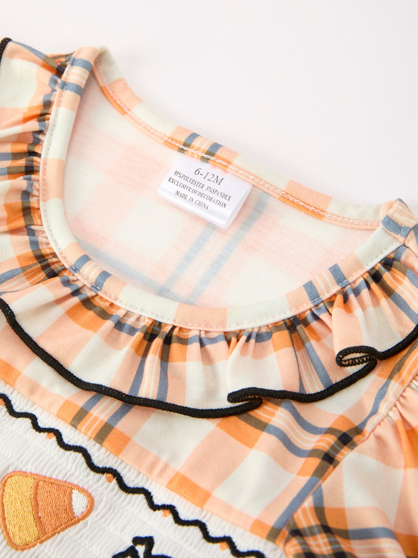 Close-up of a children's top with a peach, orange, and navy plaid pattern, featuring a ruffled neckline with black trim and a candy corn embroidery on the front.