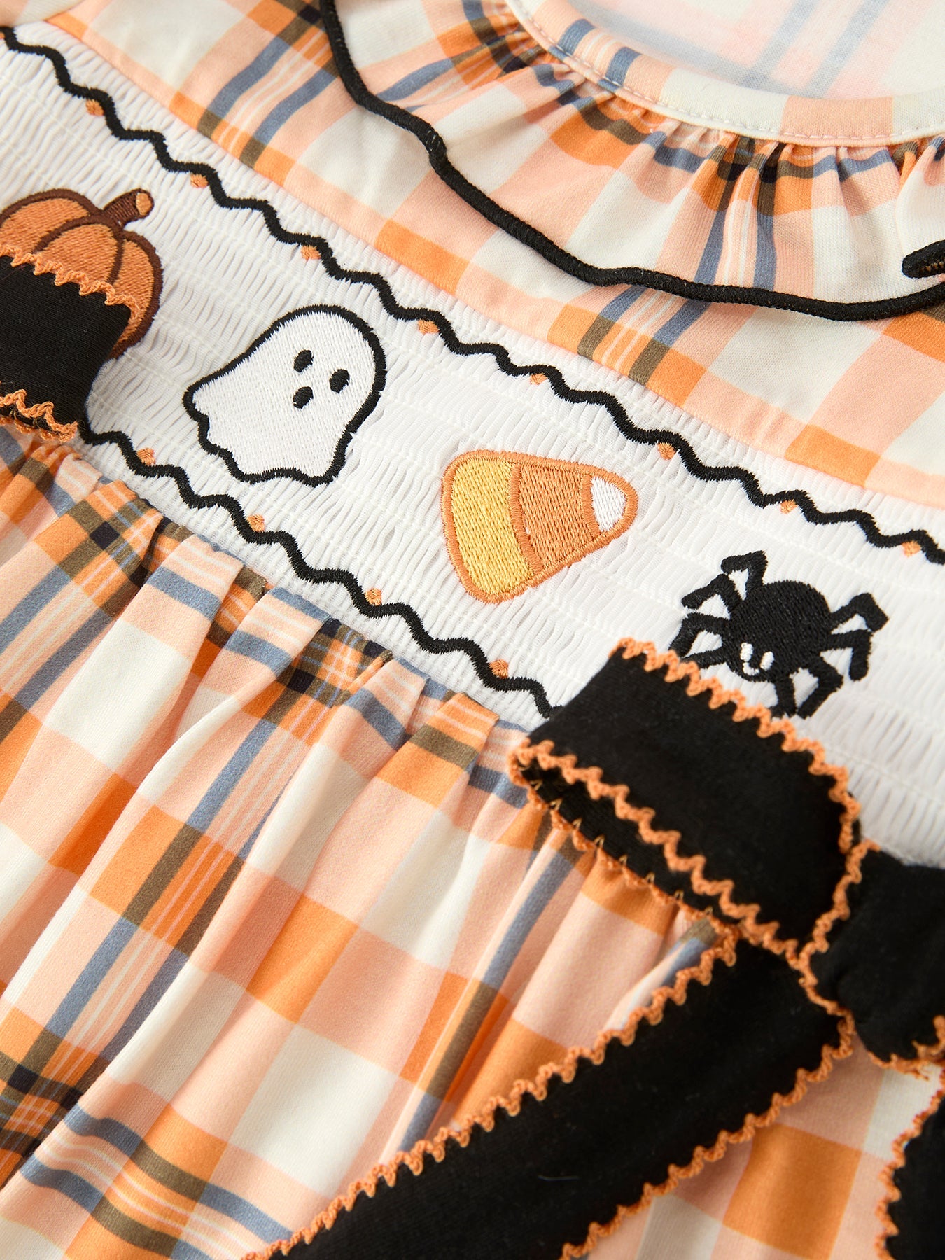 Close-up of a Halloween-themed dress featuring an orange, white, and gray plaid pattern, a smocked white band with embroidered ghosts, candy corn, a spider, and a pumpkin, and black ruffled trim with orange edging.