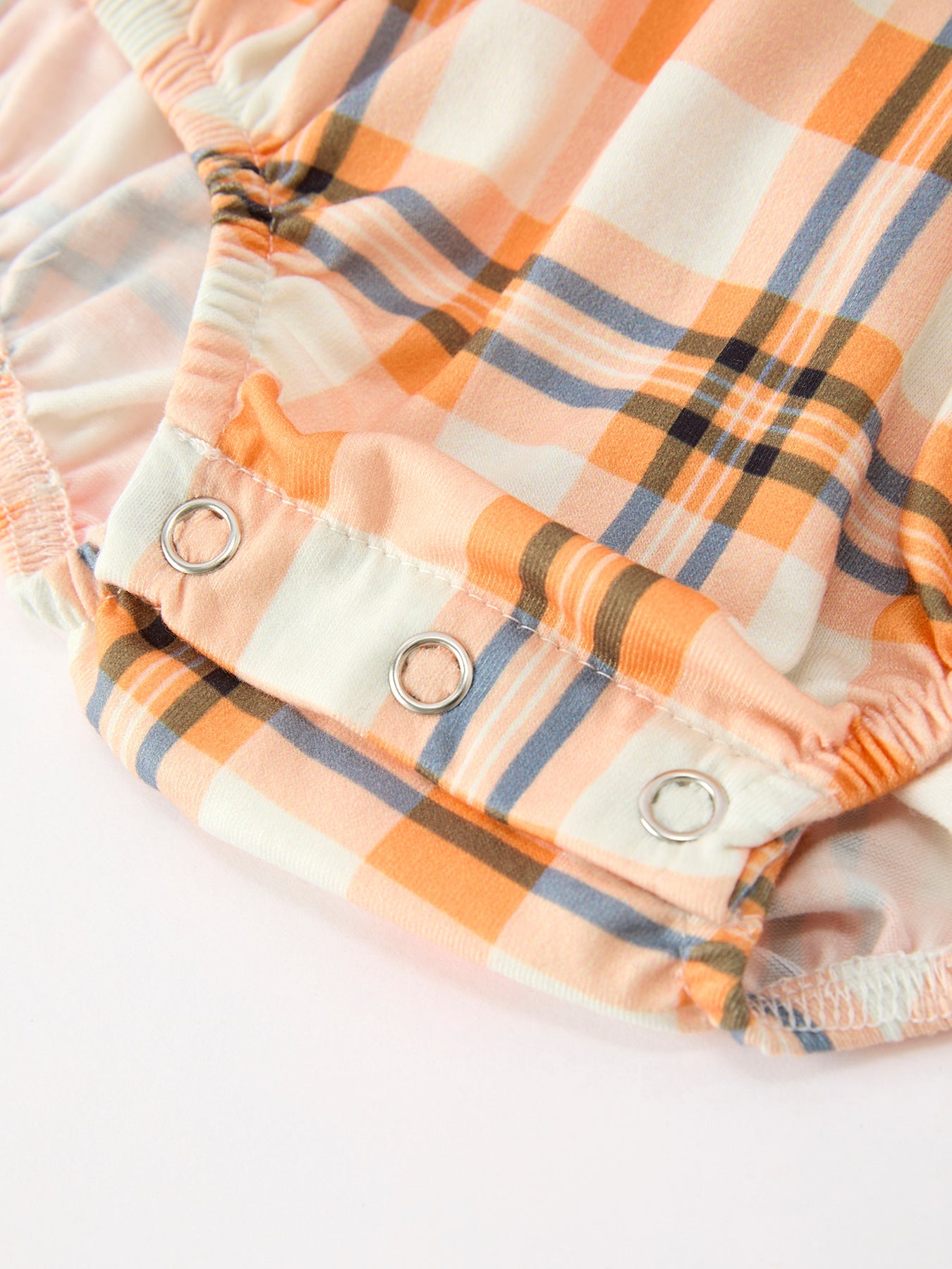 Close-up of a plaid fabric garment in peach, cream, and blue-gray, with three metal snap closures visible on the front.