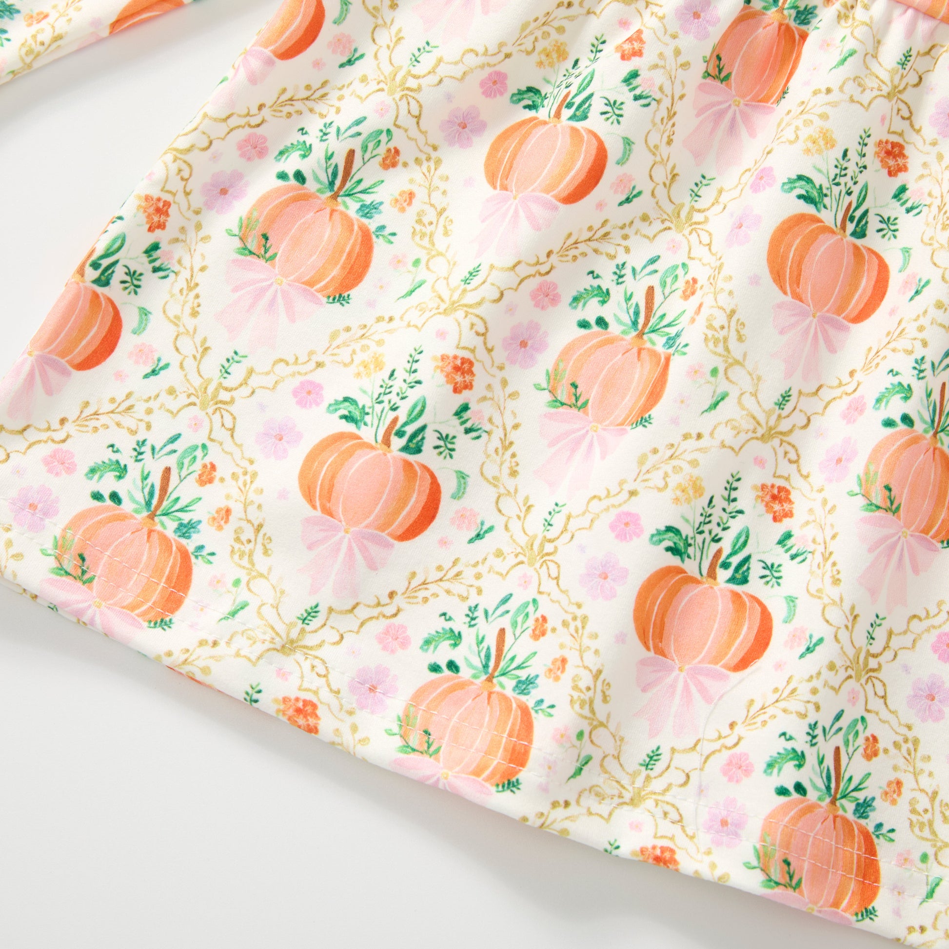 A close-up of a fabric surface with an allover pumpkin print on a white background, featuring orange pumpkins, green leaves, pink bows, and gold chain motifs.