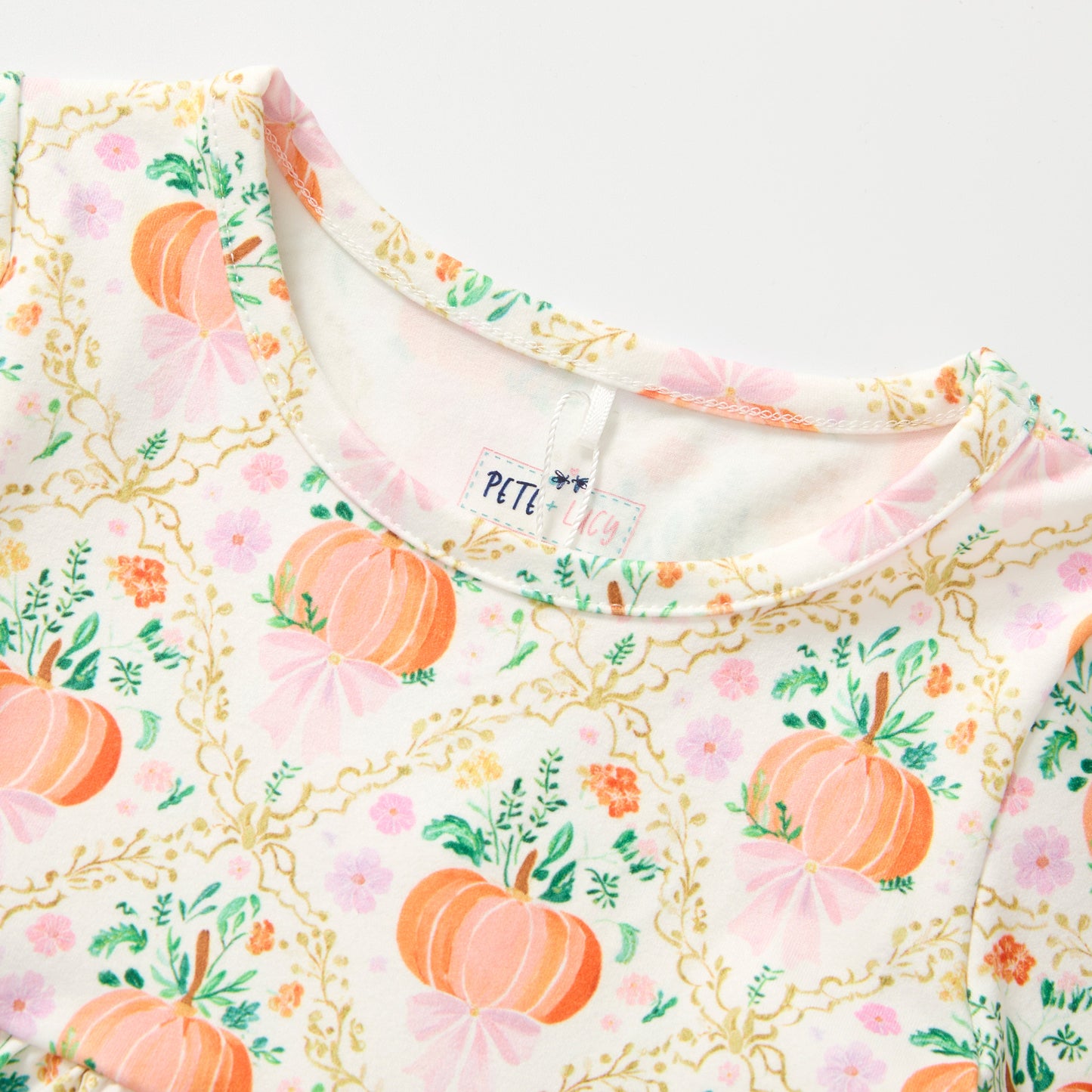 Close-up of a children's long-sleeve top featuring an autumn pumpkin and floral print on a white base. The pattern includes orange pumpkins, pink bows, green leaves, and gold vine details.