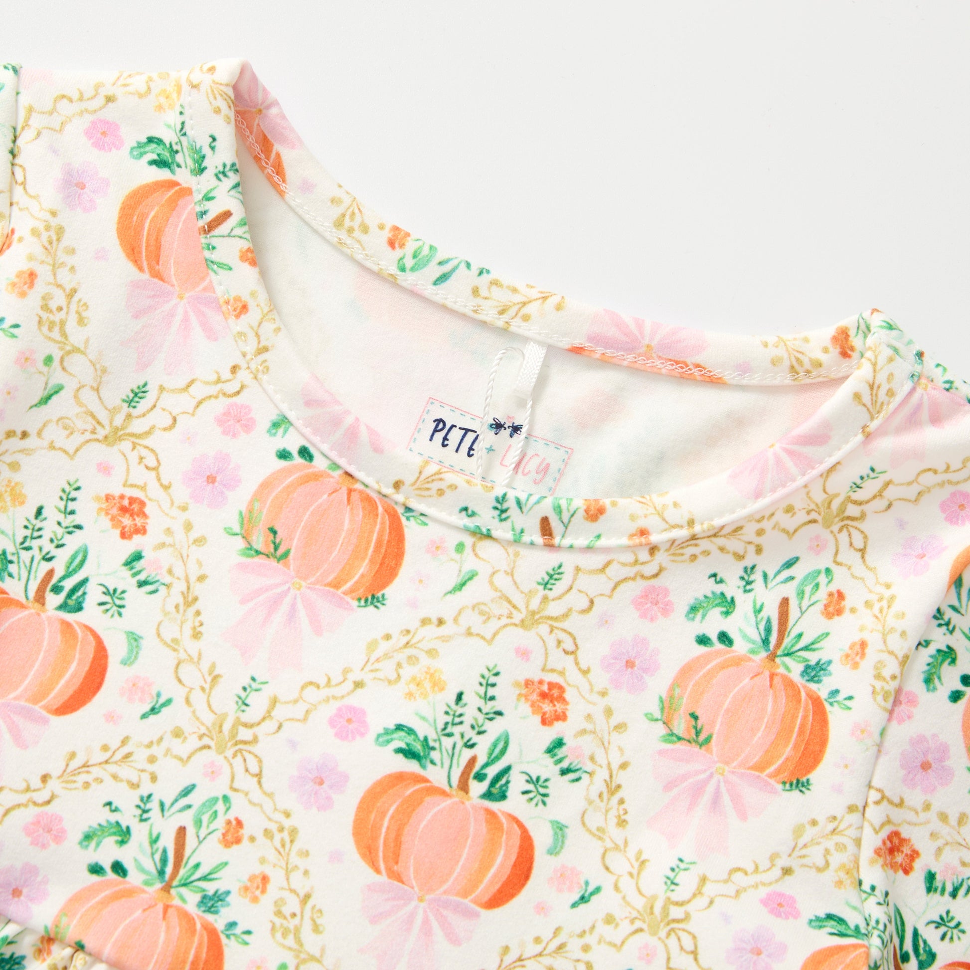 Close-up of a children's long-sleeve top featuring an autumn pumpkin and floral print on a white base. The pattern includes orange pumpkins, pink bows, green leaves, and gold vine details.