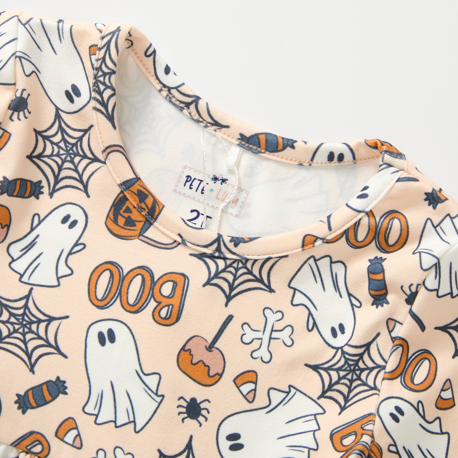 A close-up of a children's long-sleeve top with a Halloween print on a light peach background. The pattern features white ghosts, black spiderwebs, orange pumpkins, candy corn, and the word 'Boo' in orange.