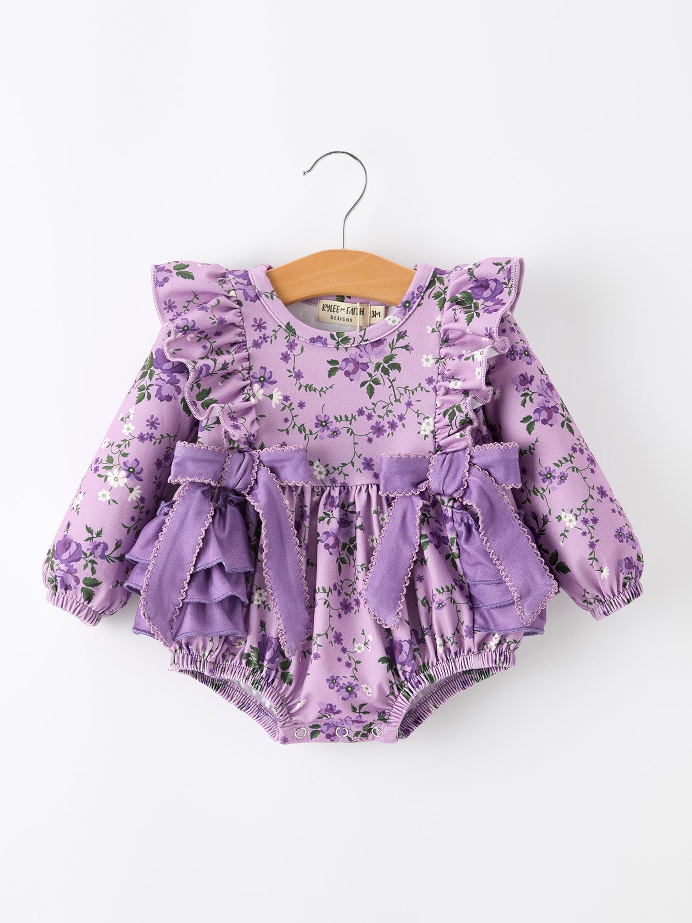 A baby girl's long-sleeve romper in a light purple floral print, featuring ruffled shoulders, a matching ruffled waist bow, and elasticized bloomers, hanging on a wooden hanger against a plain white background.
