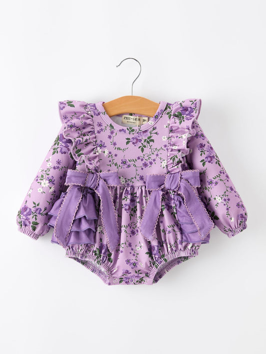 A baby girl's long-sleeve romper in a light purple floral print, featuring ruffled shoulders, a matching ruffled waist bow, and elasticized bloomers, hanging on a wooden hanger against a plain white background.