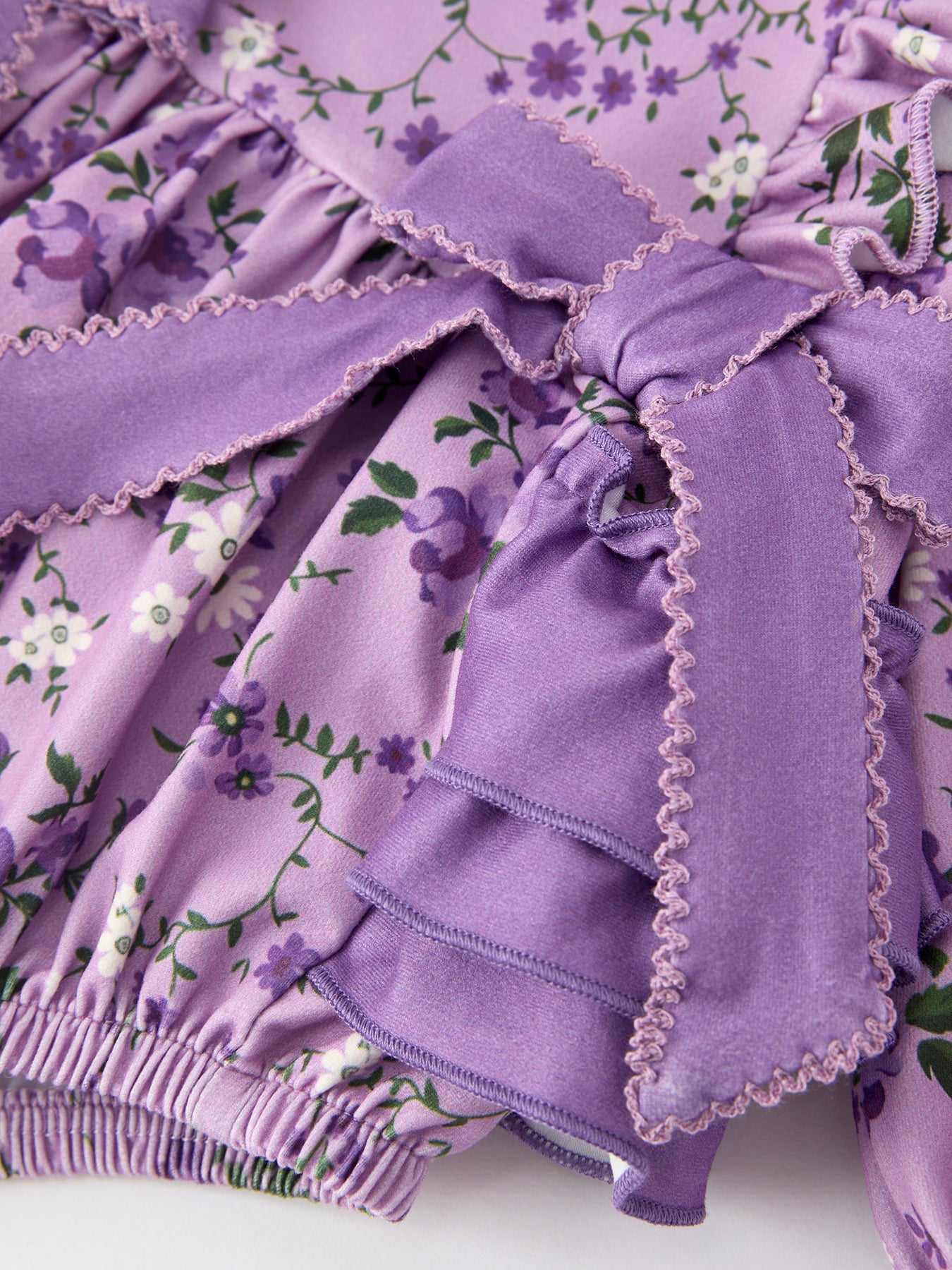 Close-up of a lavender-purple floral print garment with a large ruffled bow and matching elasticized hem, featuring small purple and white flowers with green leaves.