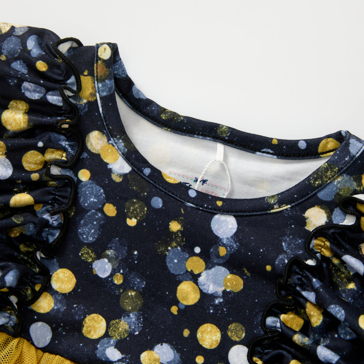 Close-up of a children's top with a navy blue base, covered in a pattern of gold, white, and gray glittery polka dots, featuring ruffled sleeves and a crew neckline.