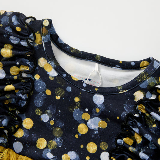 Close-up of a children's top with a navy blue base, covered in a pattern of gold, white, and gray glittery polka dots, featuring ruffled sleeves and a crew neckline.