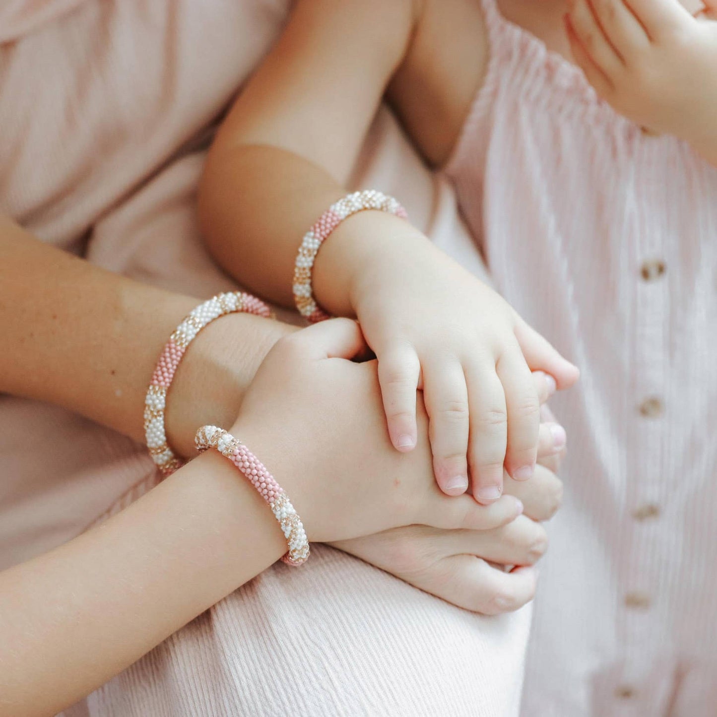 Two pairs of hands wearing pink beaded bracelets on a soft pink background.