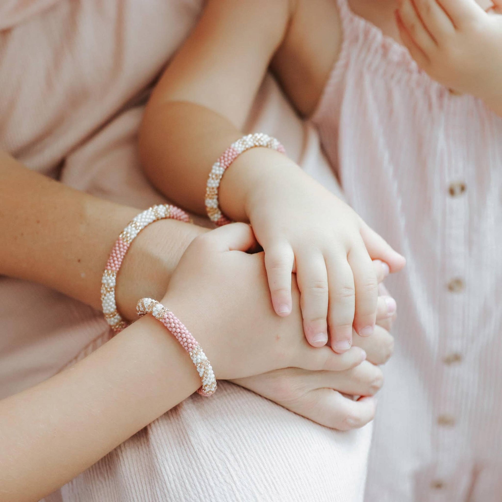 Two pairs of hands wearing pink beaded bracelets on a soft pink background.
