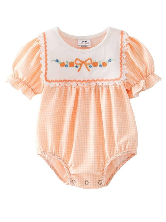 A baby romper in a soft pumpkin orange and white gingham pattern, featuring a white yoke with embroidered details of pumpkins, blue flowers and a bow, with short elastic puff sleeves.