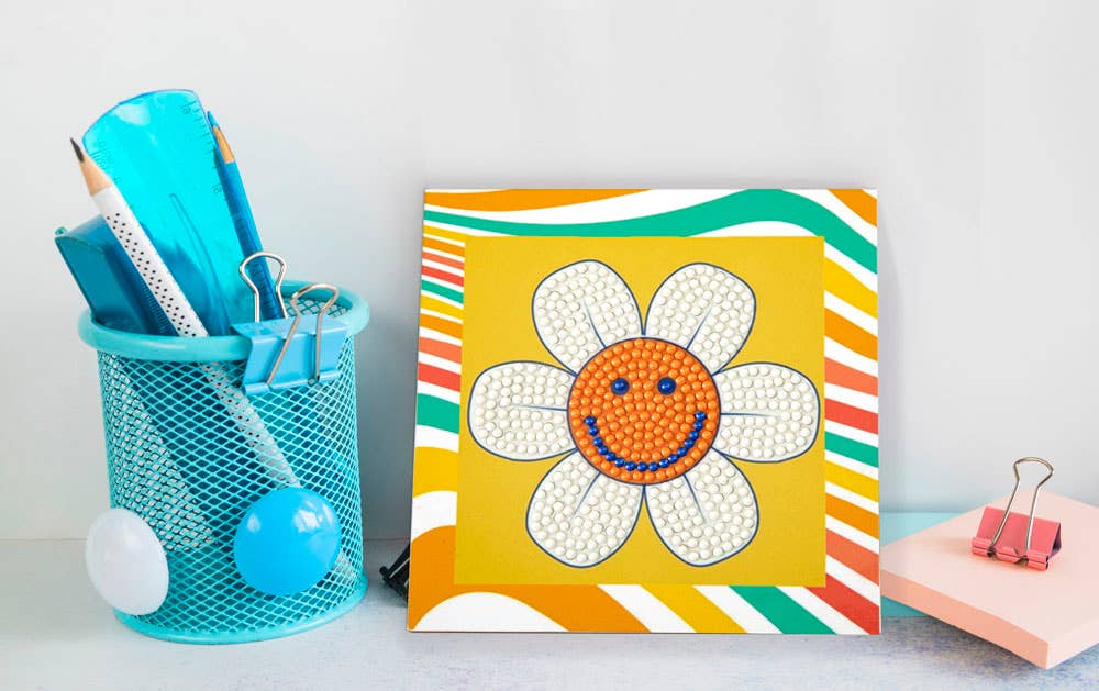 Colorful flower-shaped art piece with a smiley face on a yellow background, next to a blue mesh pencil holder with stationery items.