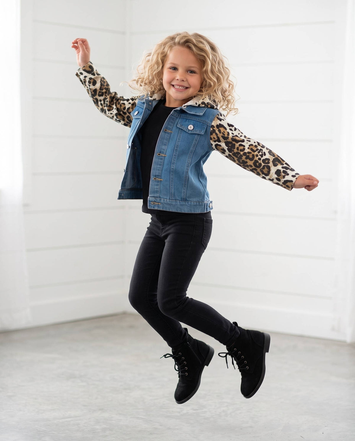 A young girl with curly blonde hair is captured mid-jump in a bright, white-paneled room. She is wearing a denim jacket with leopard-print sleeves, a black top, black pants, and black lace-up boots.