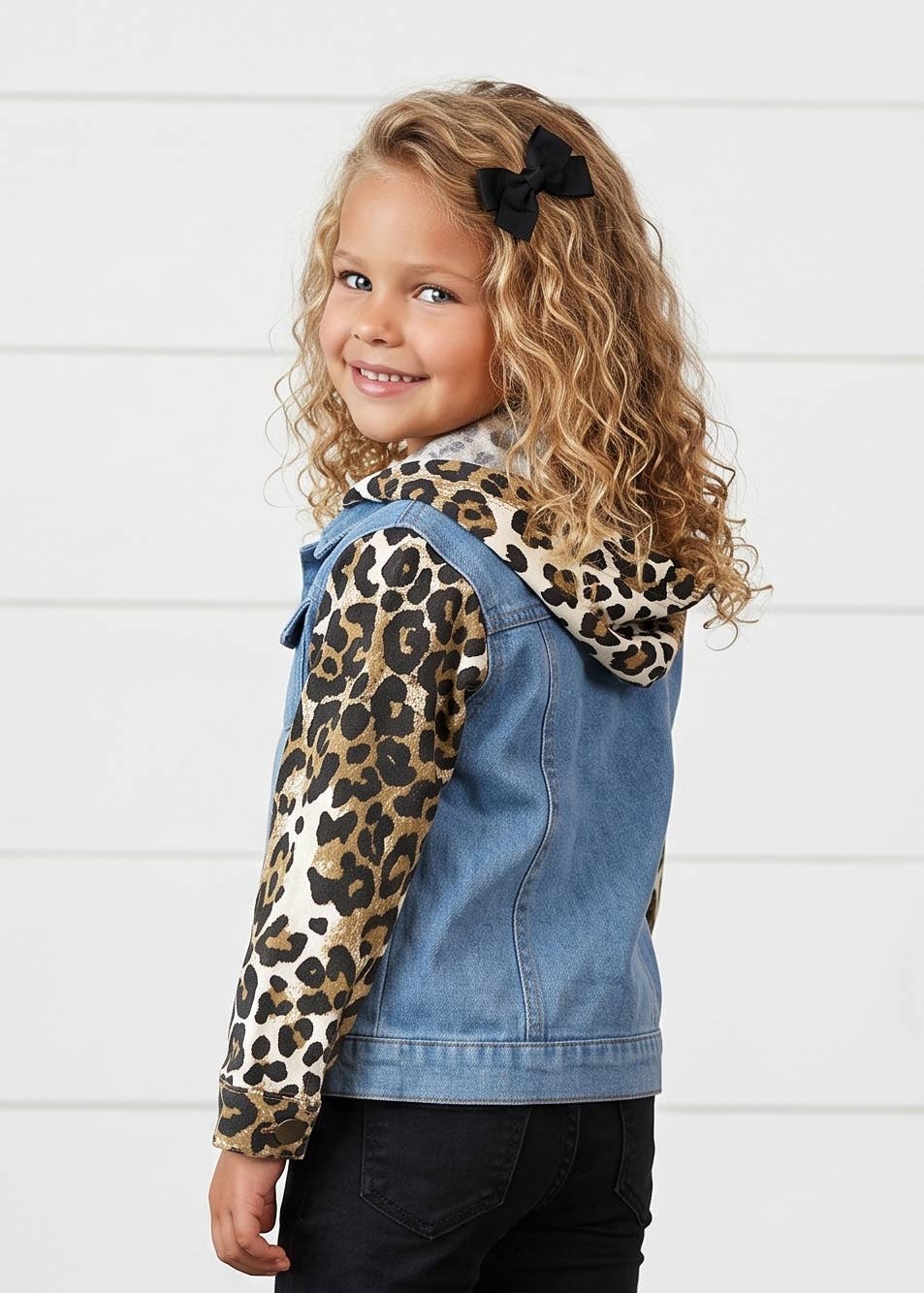 A young girl with curly blonde hair wearing a blue denim hooded jacket with leopard-print sleeves, photographed against a white paneled wall.