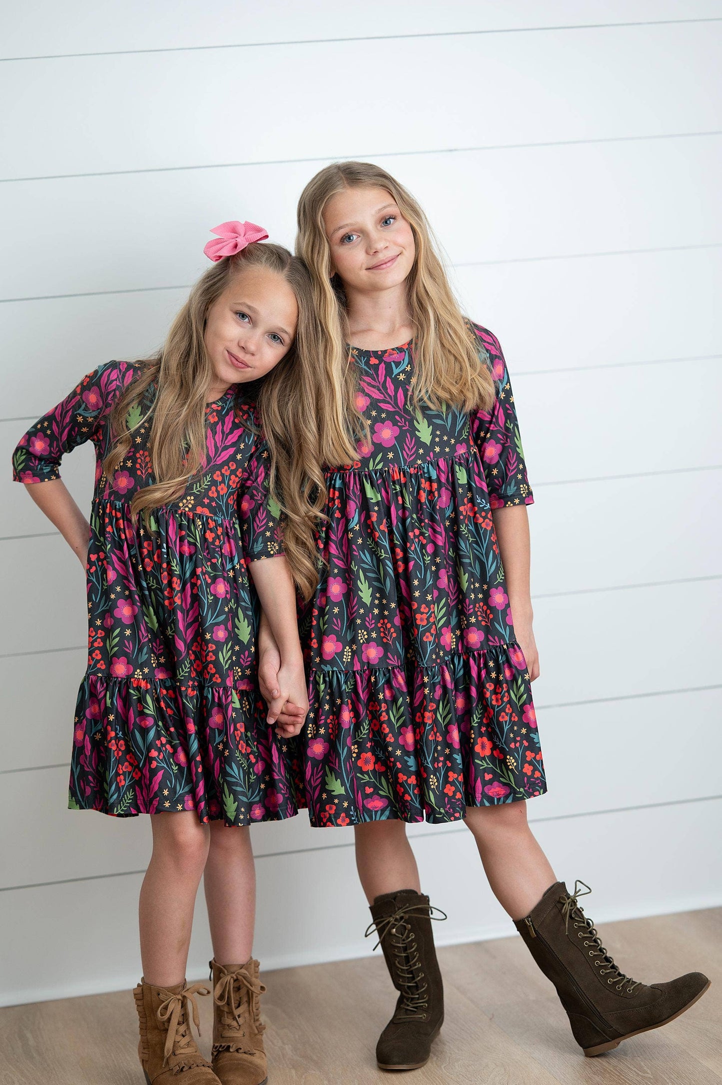 Two young girls wearing matching floral dresses against a white wooden panel background.