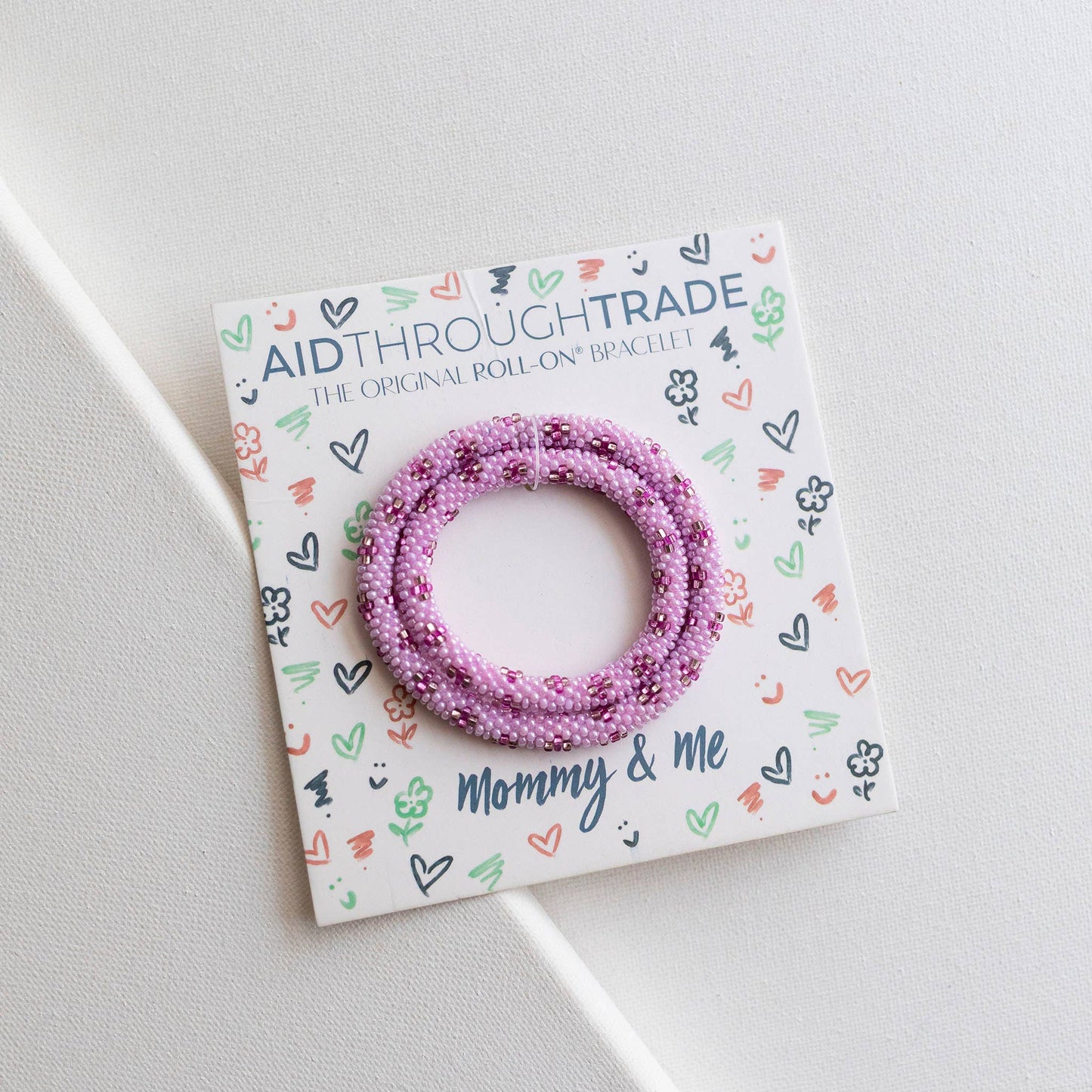 Pink beaded bracelet on a 'Aid Through Trade' branded card with decorative elements.
