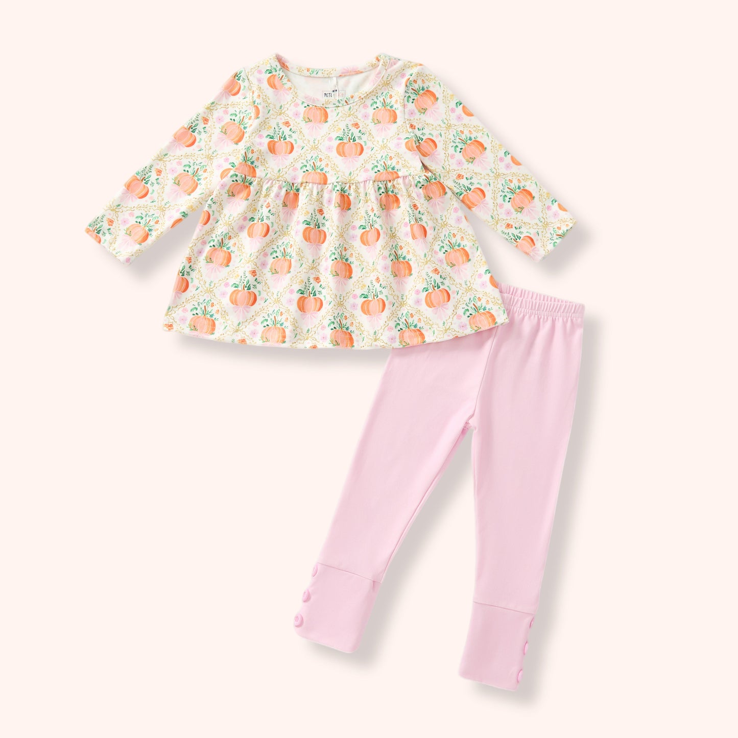 A toddler girl's two-piece outfit set featuring a long-sleeve cream-colored top with an orange and green pumpkin print, paired with light pink leggings with button cuffs.